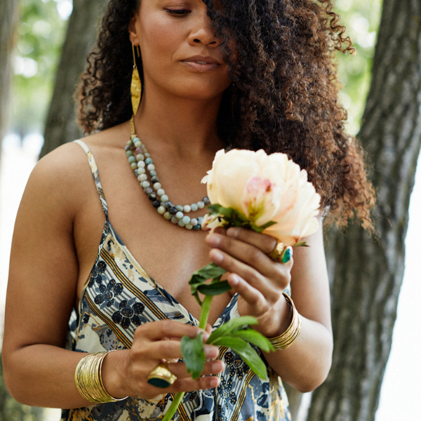 A woman outdoors among trees, wearing a patterned camisole dress and layered gold jewelry, holding a pale peach peony flower.
