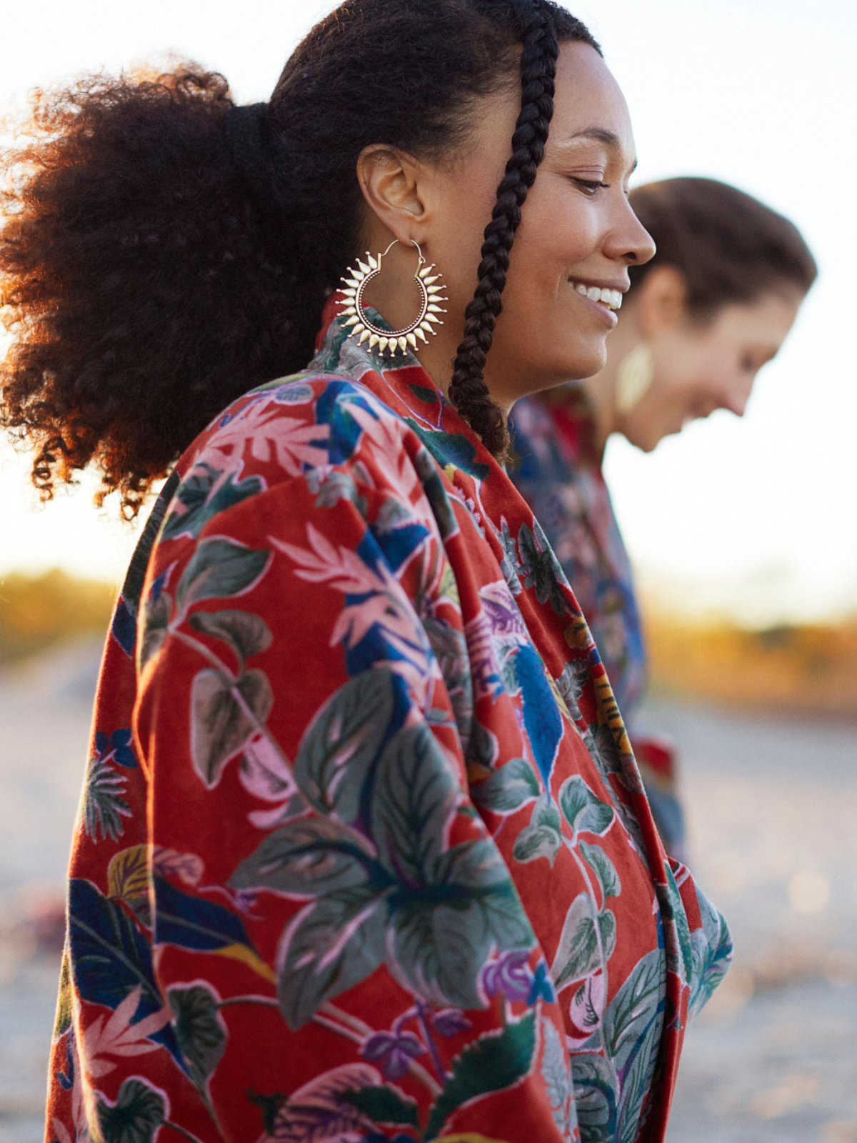 A woman in profile wearing a red floral kimono-style jacket with green, blue, and pink botanical patterns, paired with large gold hoop earrings, photographed outdoors with a softly blurred background.