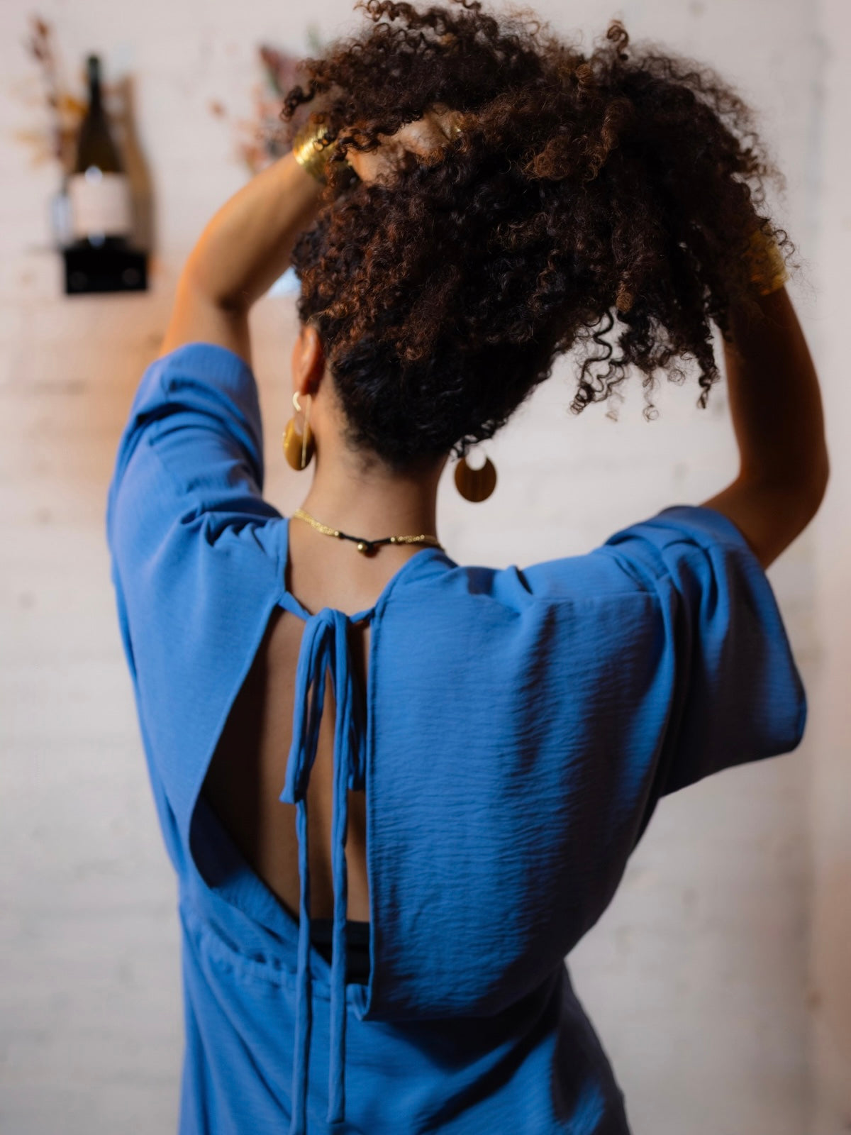 A woman viewed from behind wearing a bright blue long-sleeve top with a tie-back open back, standing against a light brick wall and touching her hair.