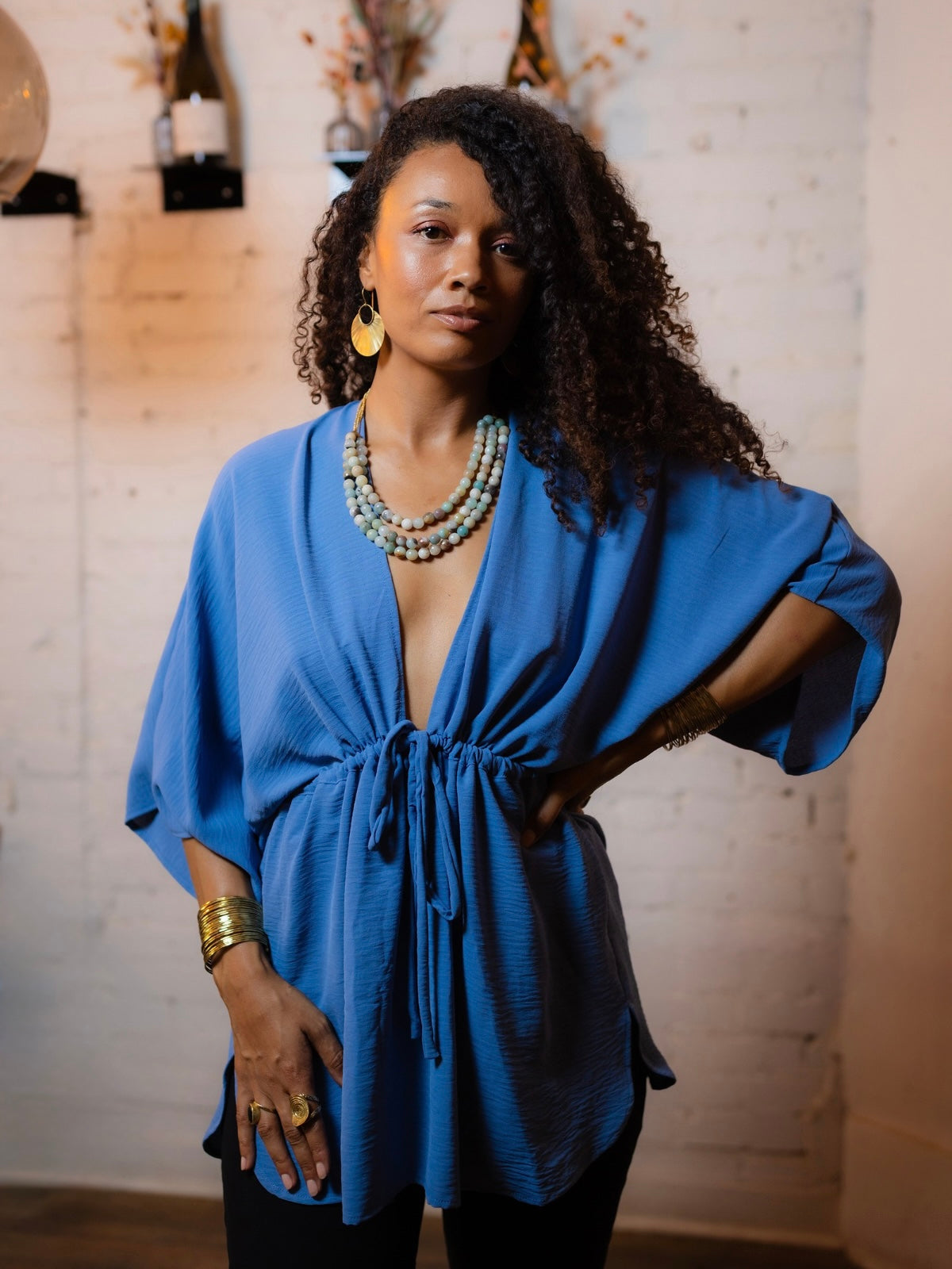 A woman with curly dark hair models a bright blue kimono-style top with a drawstring waist and wide sleeves, styled with layered beaded necklaces, gold bracelets, and rings against a rustic white brick wall background.