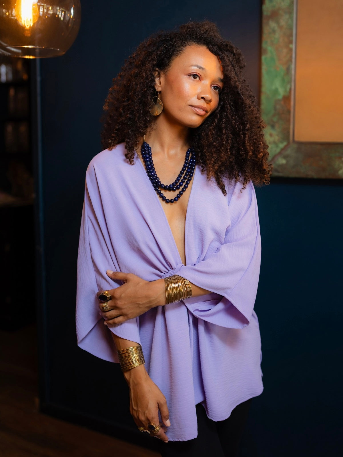 A woman with curly dark hair models a lavender wrap-style top with wide draped sleeves, styled with layered dark blue beaded necklaces and gold bracelets against a dark blue wall.