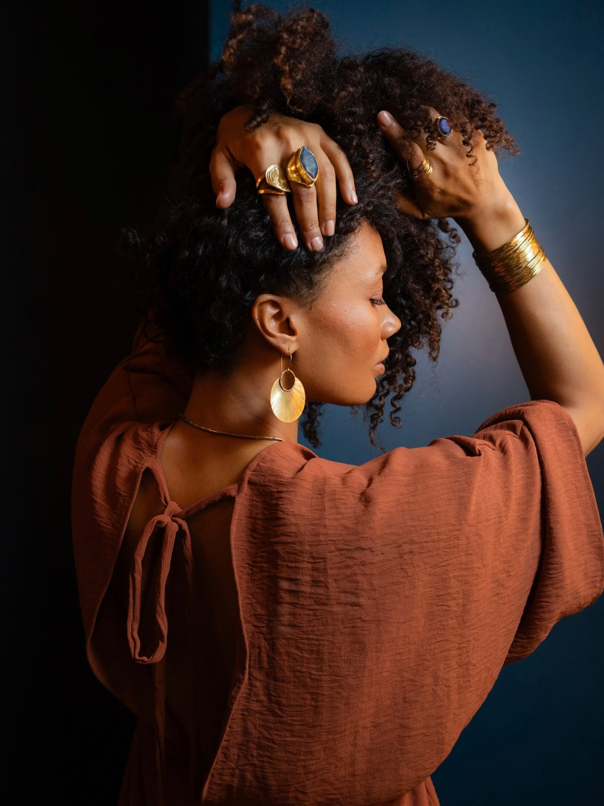 A woman with curly dark hair poses in profile wearing a rust-brown textured top and gold jewelry, including a large ring with a blue stone, hoop earrings, and multiple gold bracelets, against a dark blue studio background.