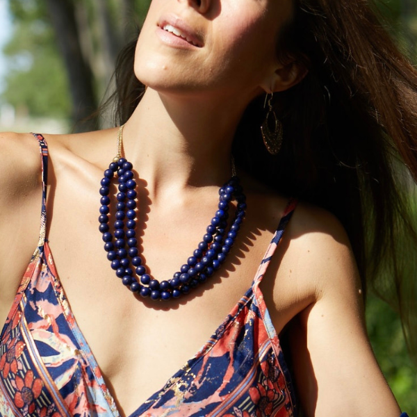 A woman outdoors wearing a layered necklace made of dark blue beads with small gold spacer beads and gold-toned chain links. She is also wearing a patterned camisole and a dangling earring.