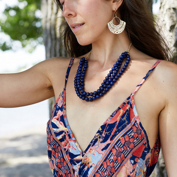 A woman outdoors wearing a dark blue beaded double-strand necklace and a patterned camisole with blue, red, and orange tones, along with a gold-tone crescent earring.