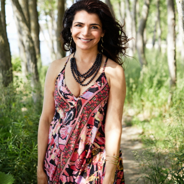 A woman standing outdoors in a wooded path wearing a sleeveless halter dress with a pink, red, black, and beige abstract print, layered with black beaded necklaces and gold bracelets.