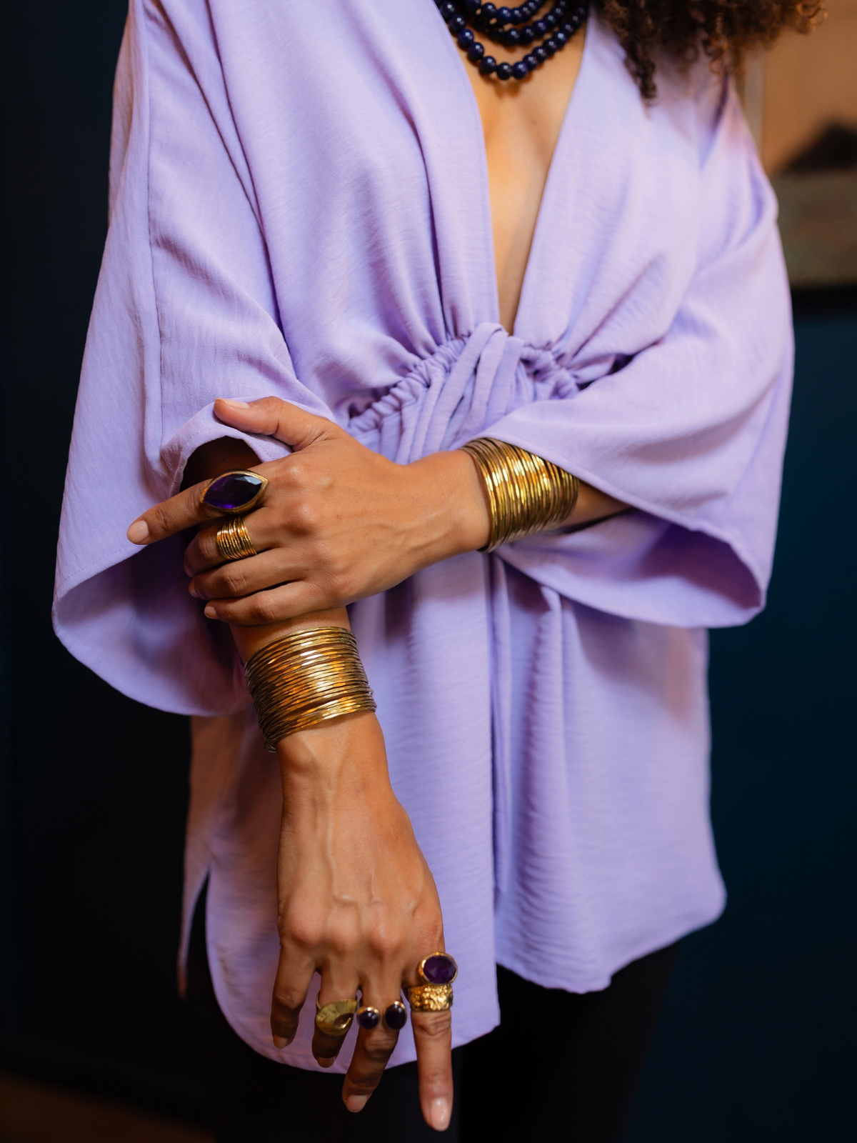 A close-up lifestyle shot of a person wearing a lavender-purple wrap-style blouse with wide sleeves, accessorized with multiple gold bracelets and rings, against a dark background.