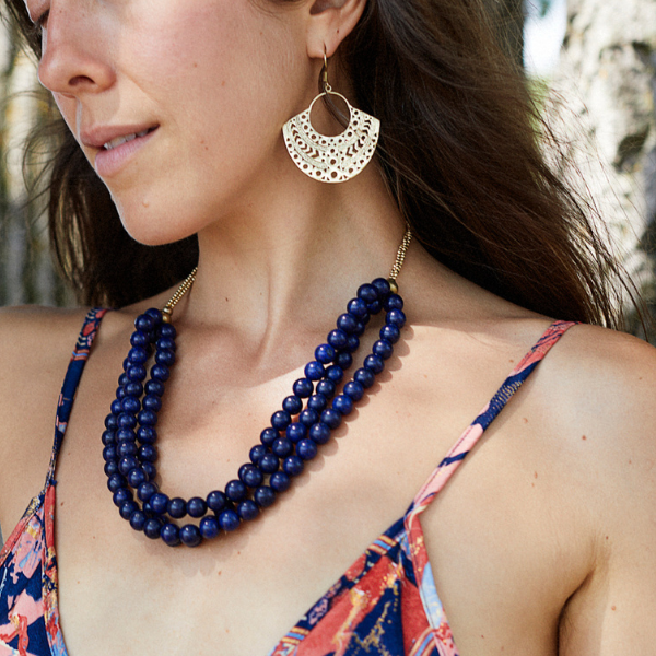 A close-up lifestyle shot of a woman wearing a multi-strand navy blue beaded necklace and a matching navy and gold semi-circular cutout dangle earring, paired with a colourful patterned dress.