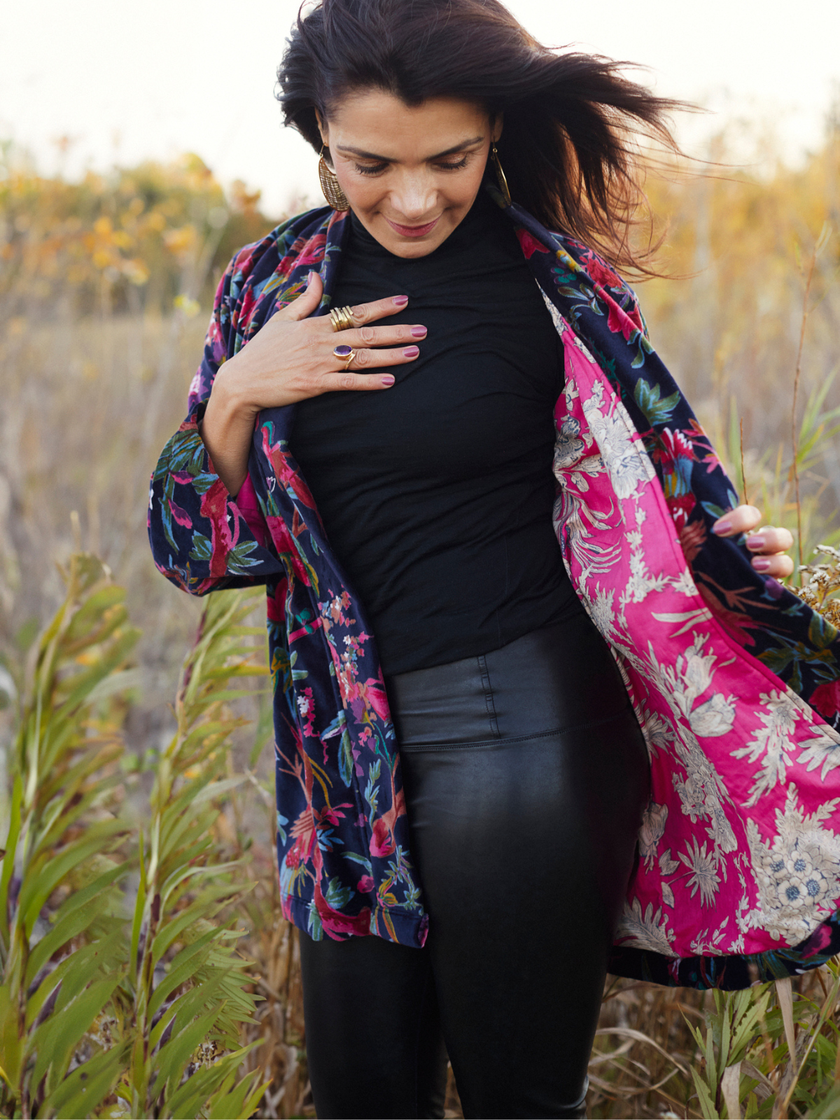 A woman standing outdoors in a field, wearing a black turtleneck and black pants, with a colorful floral kimono-style jacket draped open over her shoulders. The jacket has a navy blue outer pattern and a bright pink inner lining with white floral motifs.