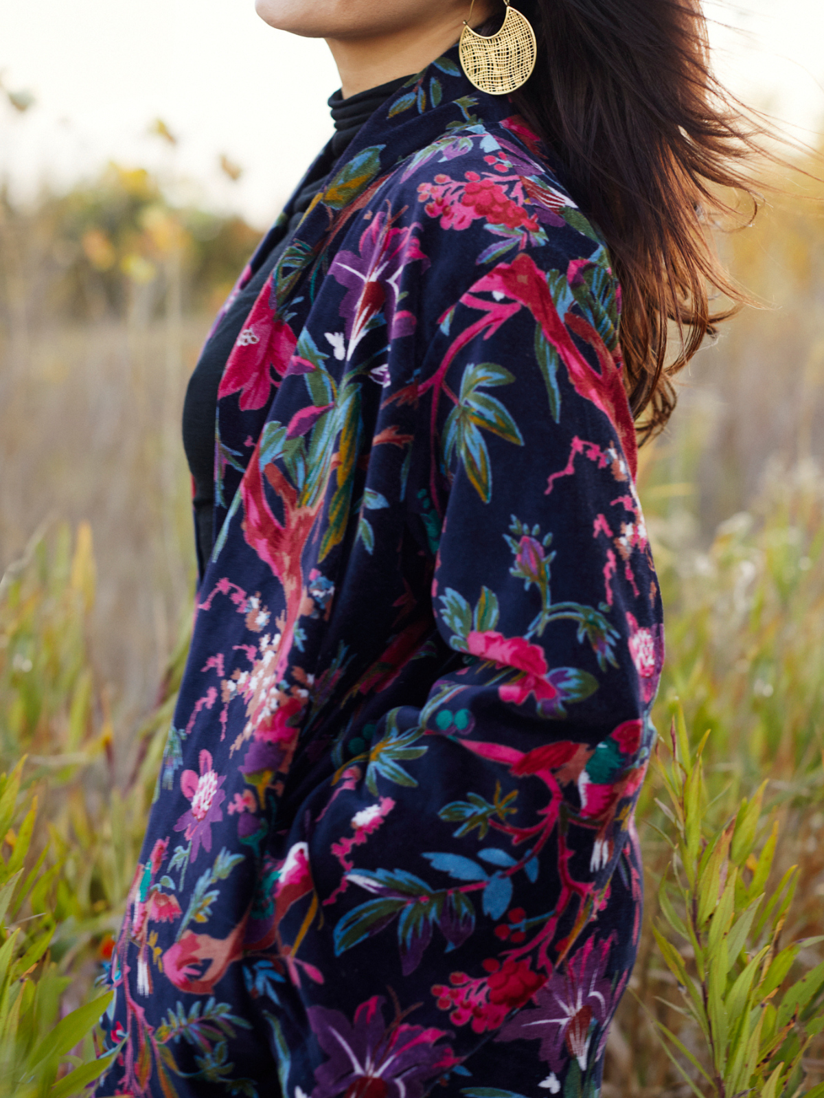 A woman wearing a dark navy floral jacket with pink, red, and green botanical patterns, photographed outdoors in a grassy field at sunset.