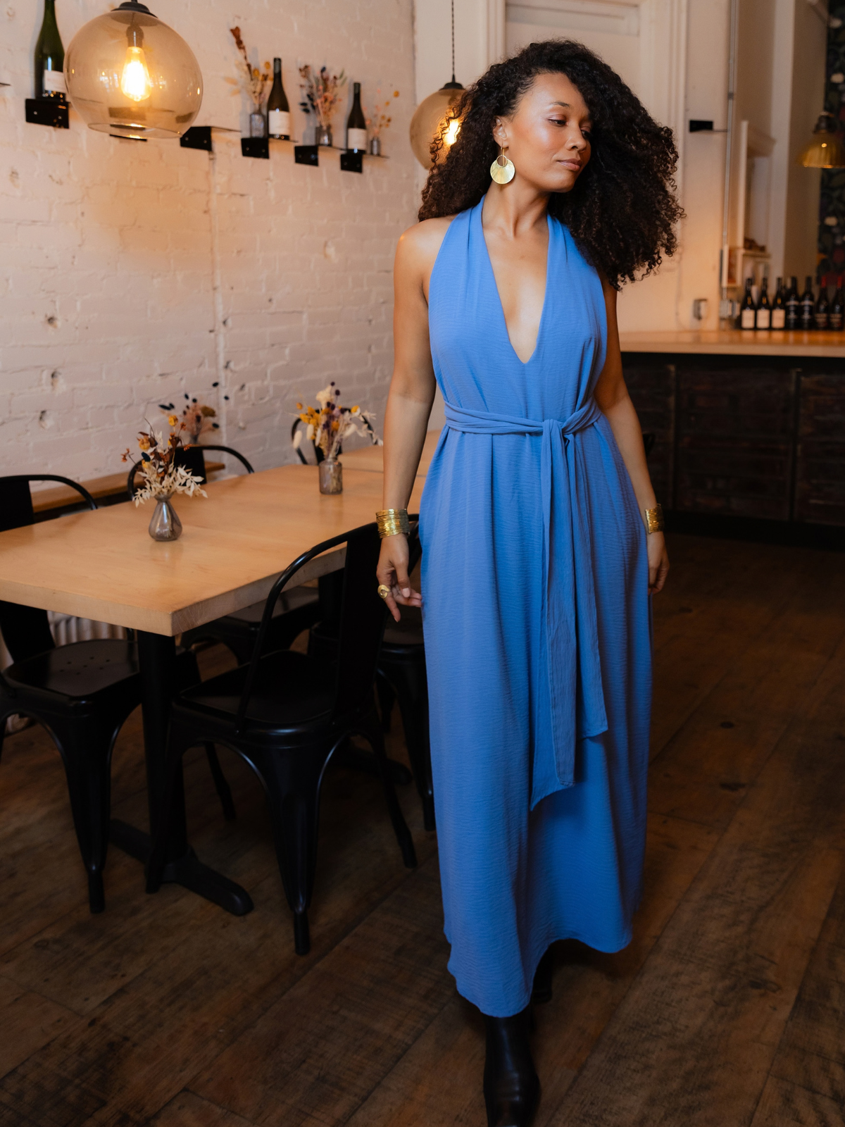 A woman with curly dark hair wearing a sleeveless, deep V-neck, blue maxi dress with a tie waist, walking in a rustic restaurant or cafe with wooden floors, black chairs, and warm pendant lights.