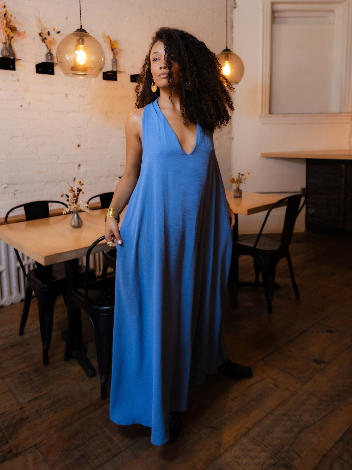 A woman with curly hair wearing a sleeveless, deep V-neck blue maxi dress stands in a rustic cafe with wooden floors, black chairs, and hanging glass pendant lights.