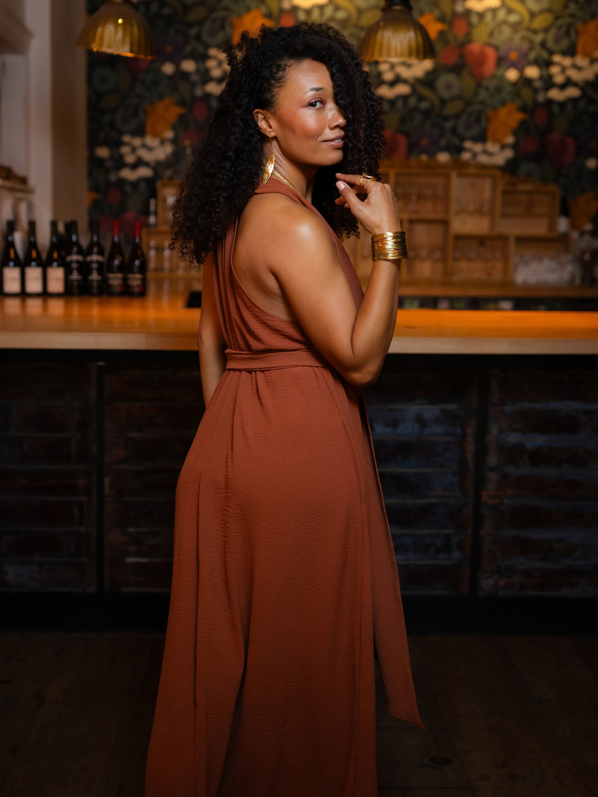 A woman with curly black hair stands in a bar setting wearing a sleeveless rust-brown maxi dress with a waist tie and gold jewelry.