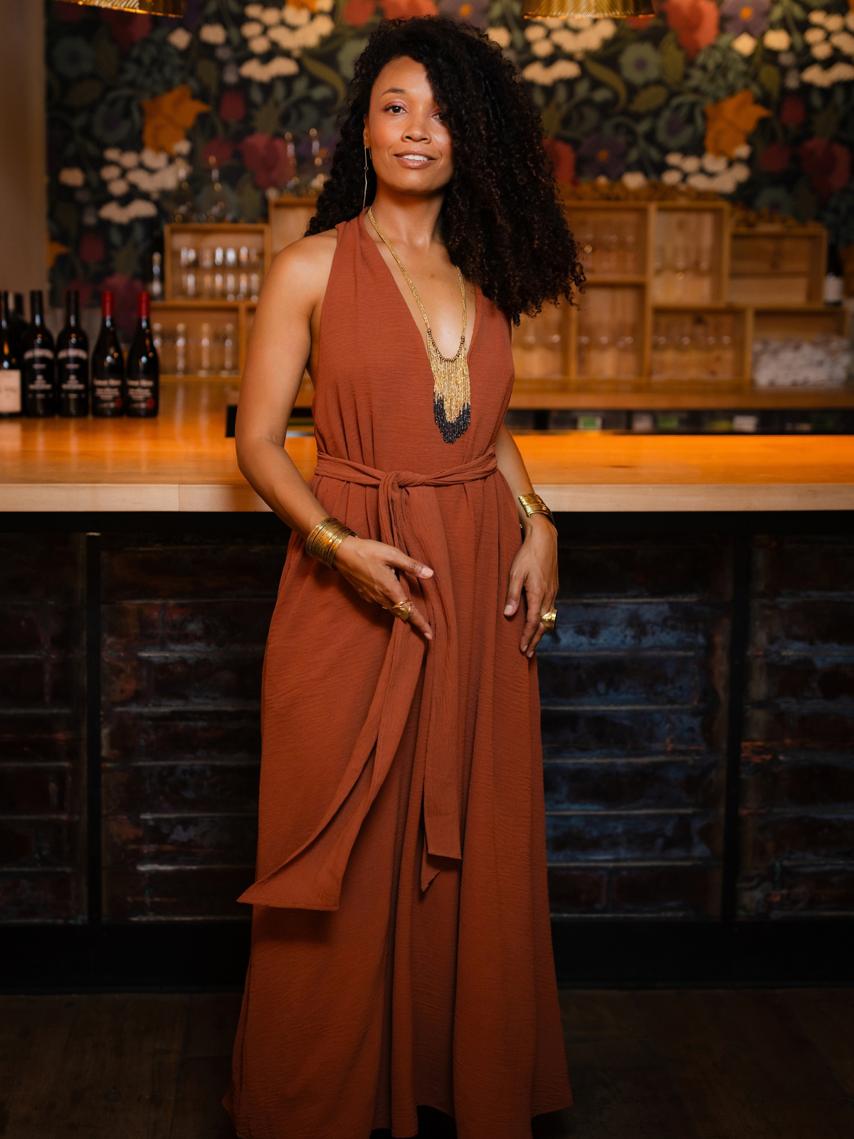 A woman with curly black hair stands in a bar setting wearing a rust-brown sleeveless maxi dress with a tie waist and layered gold jewelry.