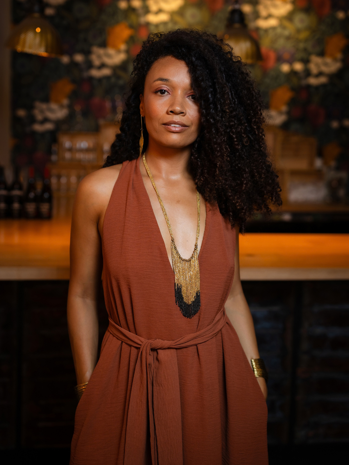 A woman with curly black hair wearing a rust-brown halter dress and a gold statement necklace, standing in a warmly lit bar or restaurant setting.