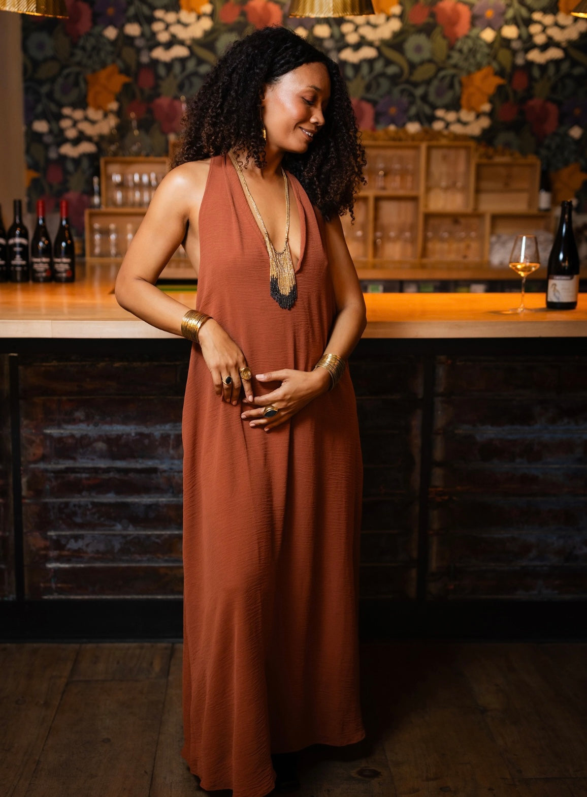 A woman with curly black hair stands in a warmly lit bar or restaurant setting wearing a sleeveless rust-orange maxi dress with a deep V-neckline. She is accessorized with gold jewelry, including a statement necklace and bracelets, and is posing with her hands clasped in front of a dark wood counter and a floral-patterned back wall.