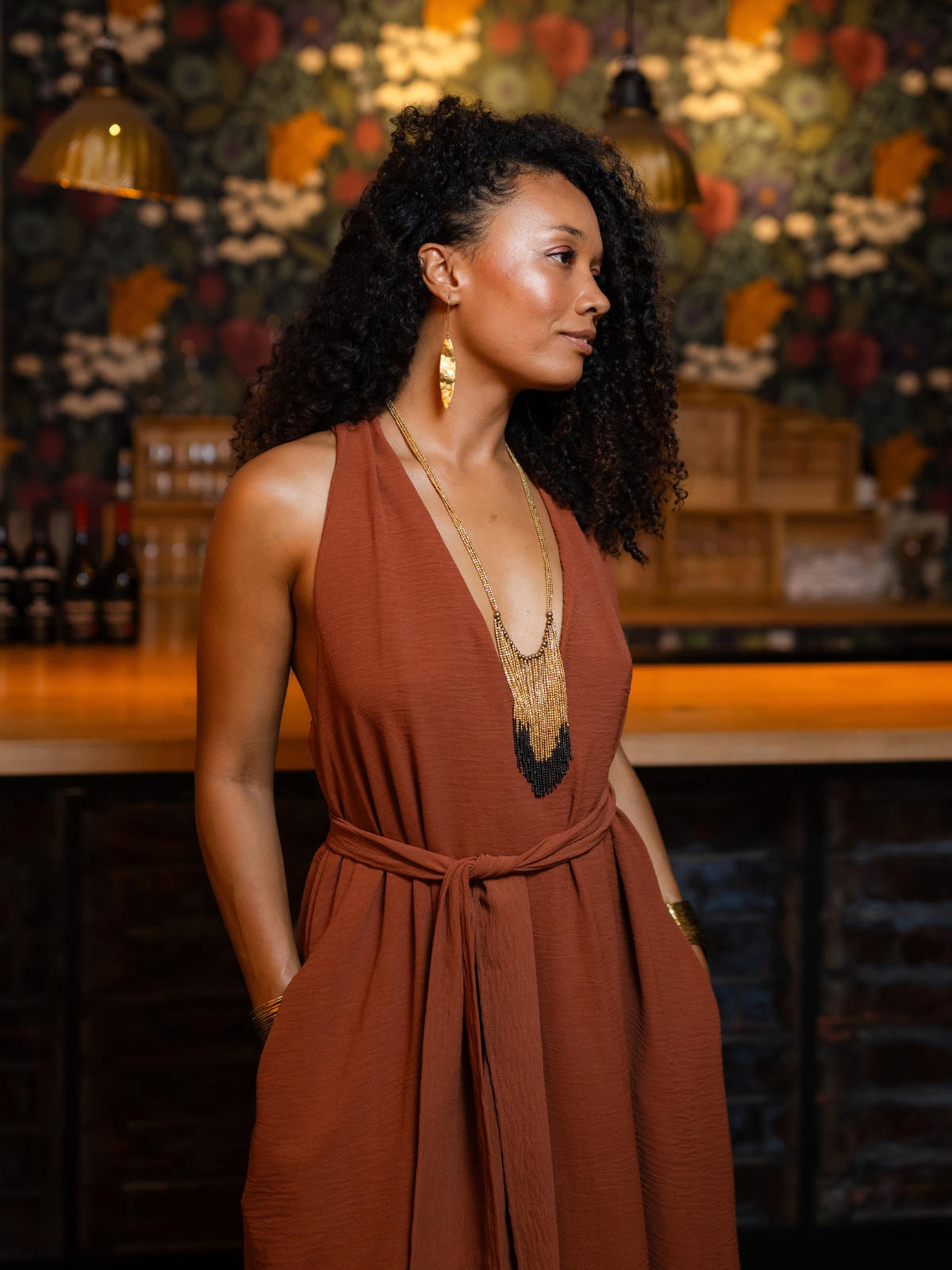 A woman with curly black hair stands in a warmly lit bar or restaurant setting, wearing a rust-brown sleeveless wrap dress with a deep V-neck and layered gold necklaces.