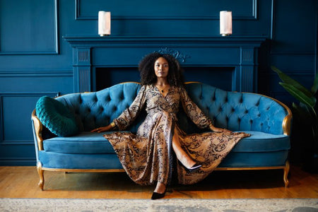 Woman in a patterned dress sitting on a blue velvet sofa against a dark blue wall.