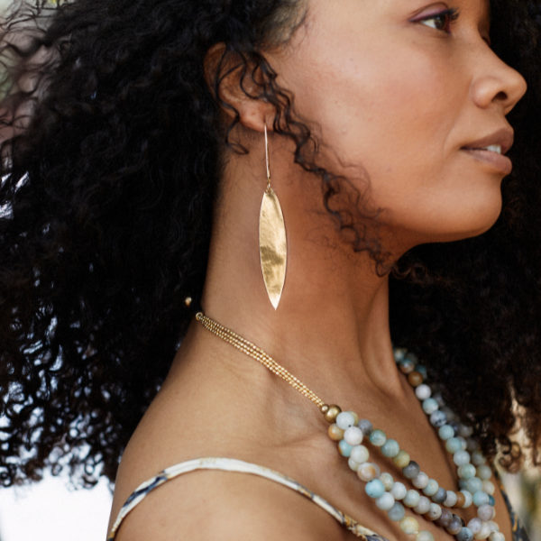 A close-up side profile of a woman with curly black hair wearing a long, hammered gold leaf-shaped dangle earring, a gold chain necklace, and layered beaded necklaces.
