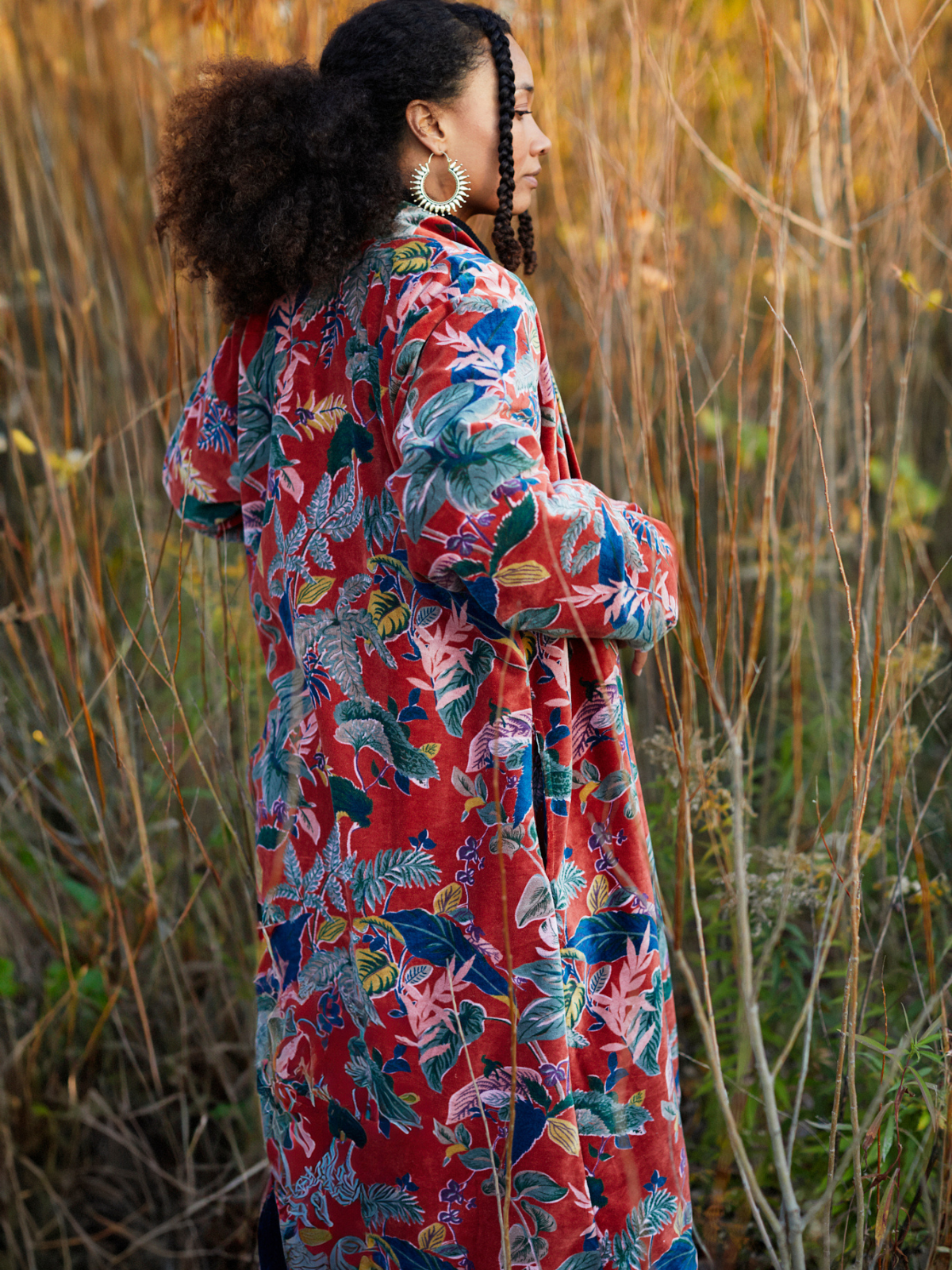 A woman wearing a long-sleeved red maxi dress with a vibrant tropical floral print in blue, green, and pink, standing in a field of dry golden grass.