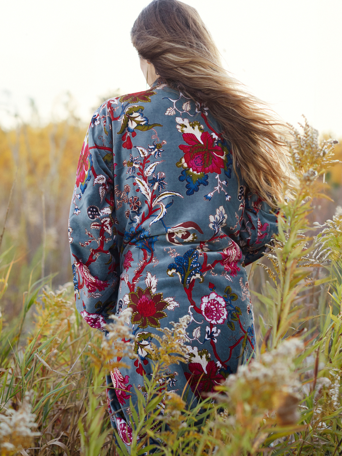 A woman viewed from behind wearing a long-sleeve blue-gray jacket with a vibrant, multicolored floral and bird pattern, standing in a field of dry grass.
