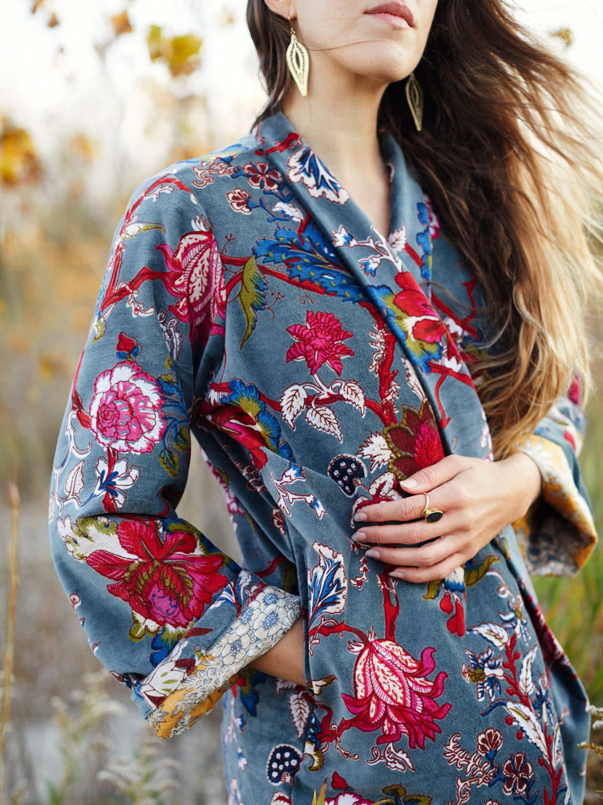 A woman wearing a blue-gray floral robe with vibrant pink, red, and green botanical patterns, photographed outdoors against a soft-focus natural background.