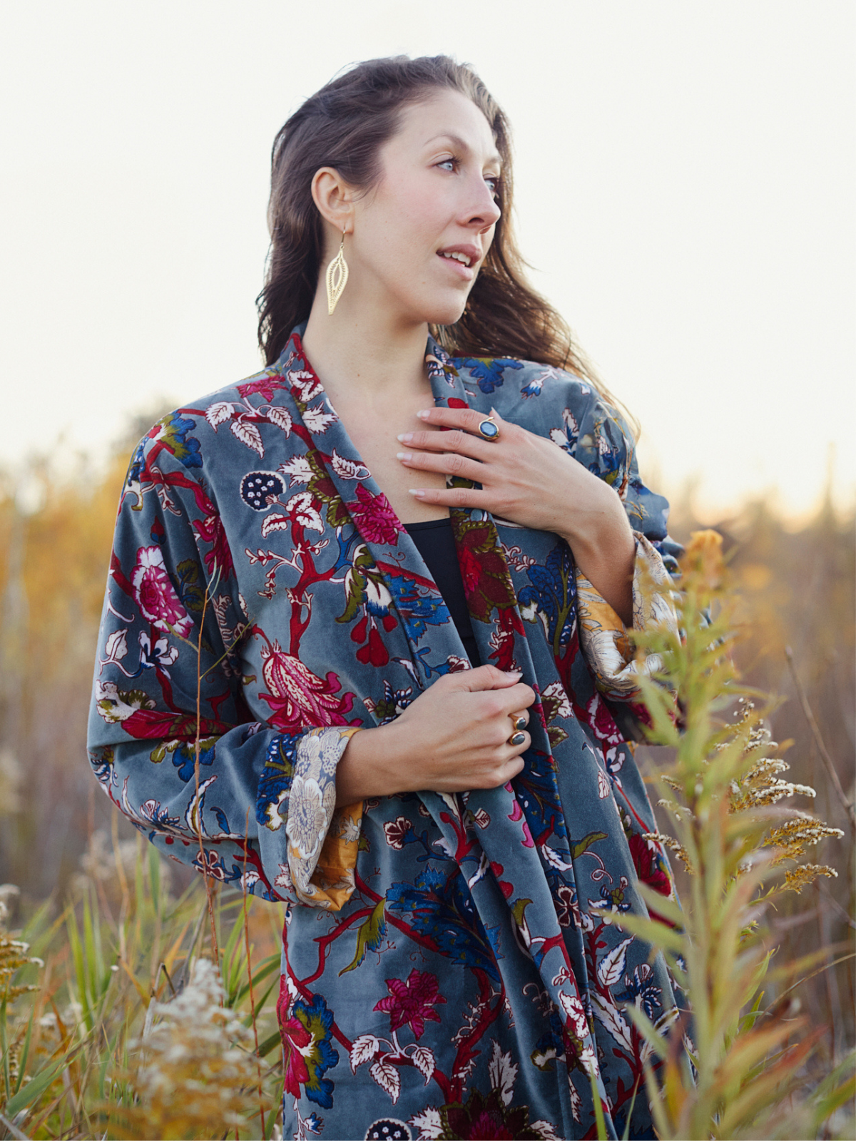 A woman wearing a blue floral kimono-style cardigan with red, pink, and green botanical patterns, standing outdoors in a field of tall grass during golden hour.