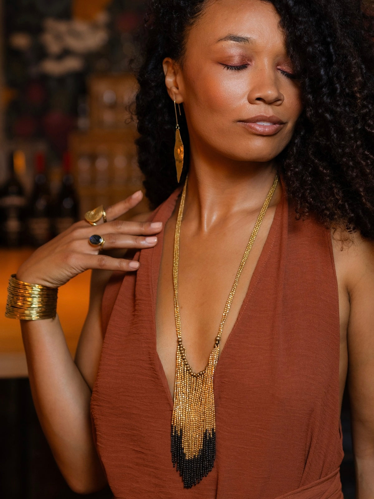 A woman wearing a long gold beaded fringe necklace and matching gold drop earrings, styled with a rust-brown halter top. She is shown in a warm, dimly lit indoor setting.