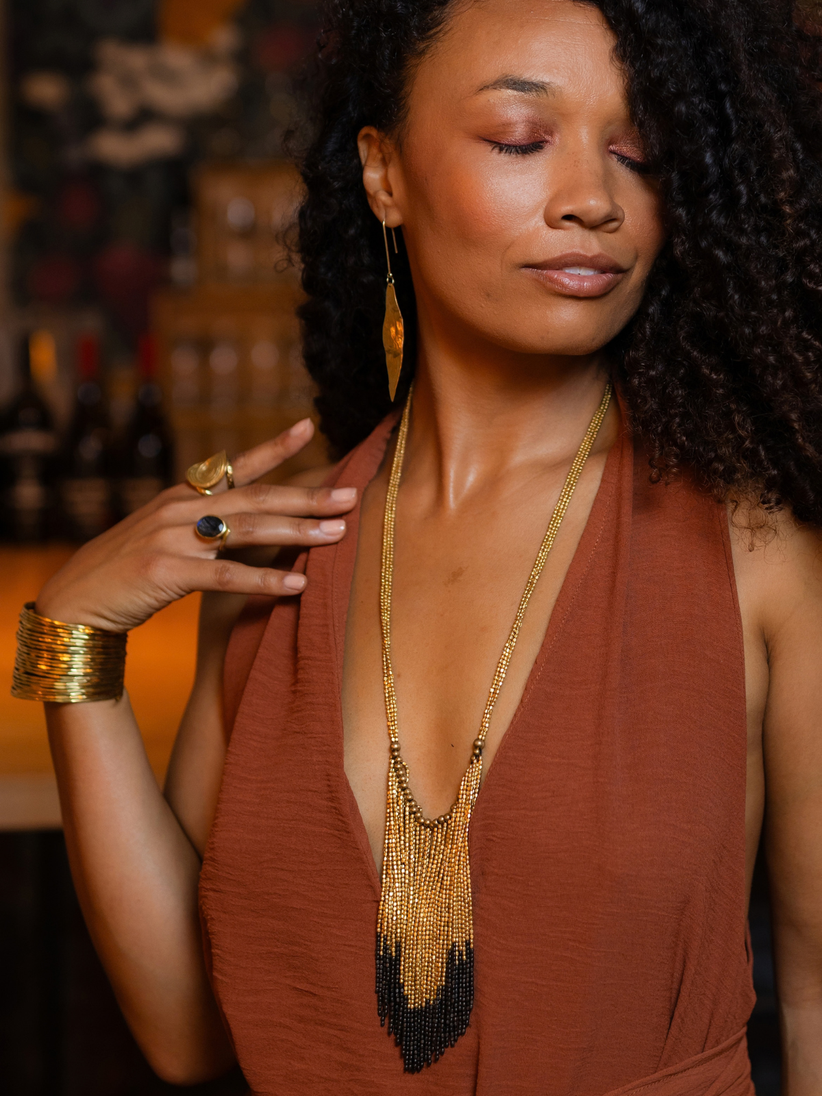 A woman with curly dark hair wearing a rust-brown halter top and a long gold beaded fringe necklace, with matching gold earrings and bracelets, posed against a warm, blurred background.