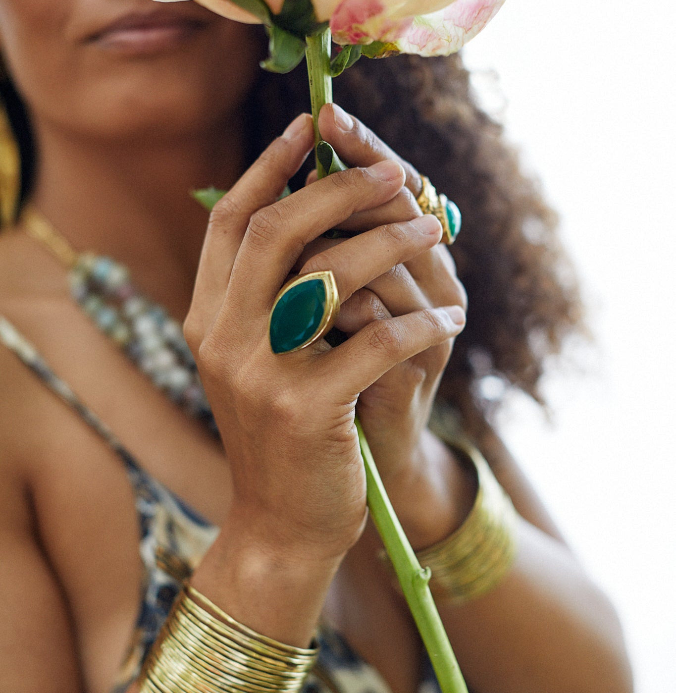 A close-up lifestyle shot of a woman holding a pale peach rose, wearing multiple gold rings and stacked gold bangles. She is wearing a patterned top, and the background is a bright, overexposed white.