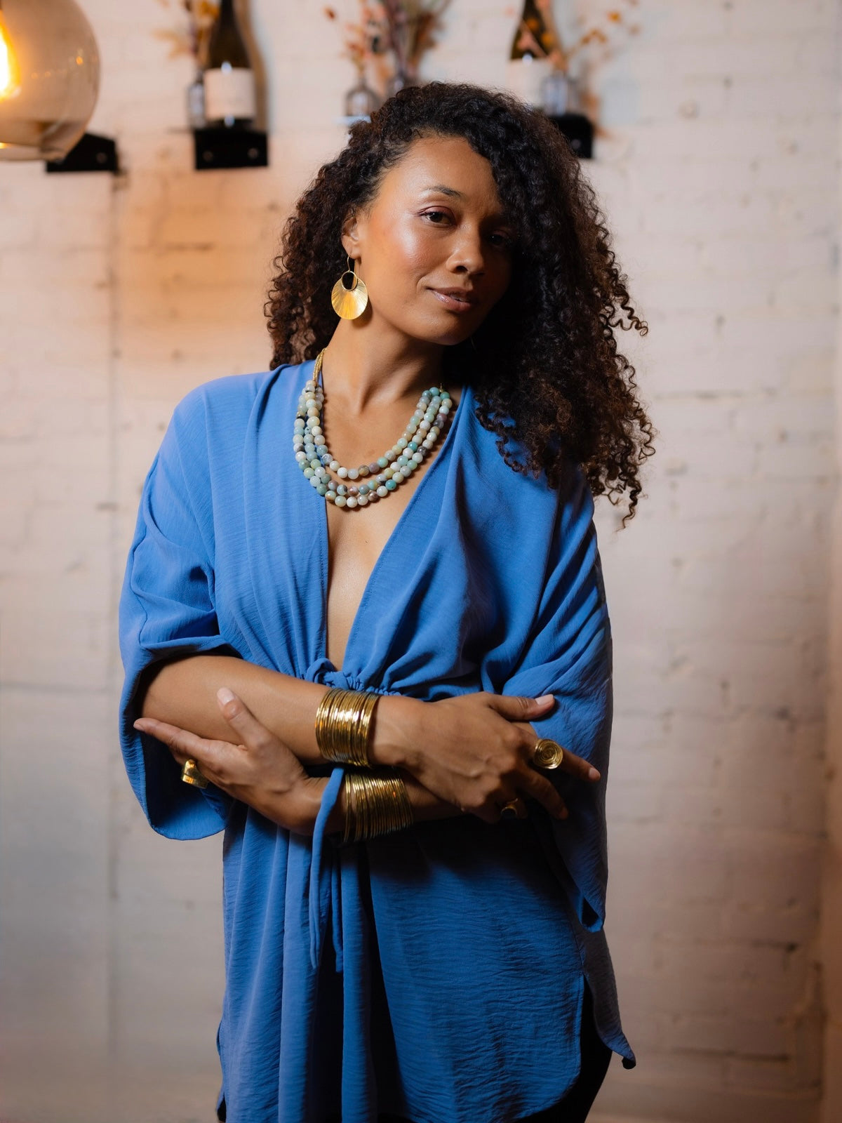 A woman with curly dark hair wearing a vibrant blue draped wrap top, layered gold bracelets, and gold earrings, posing in front of a warm-lit white brick wall.