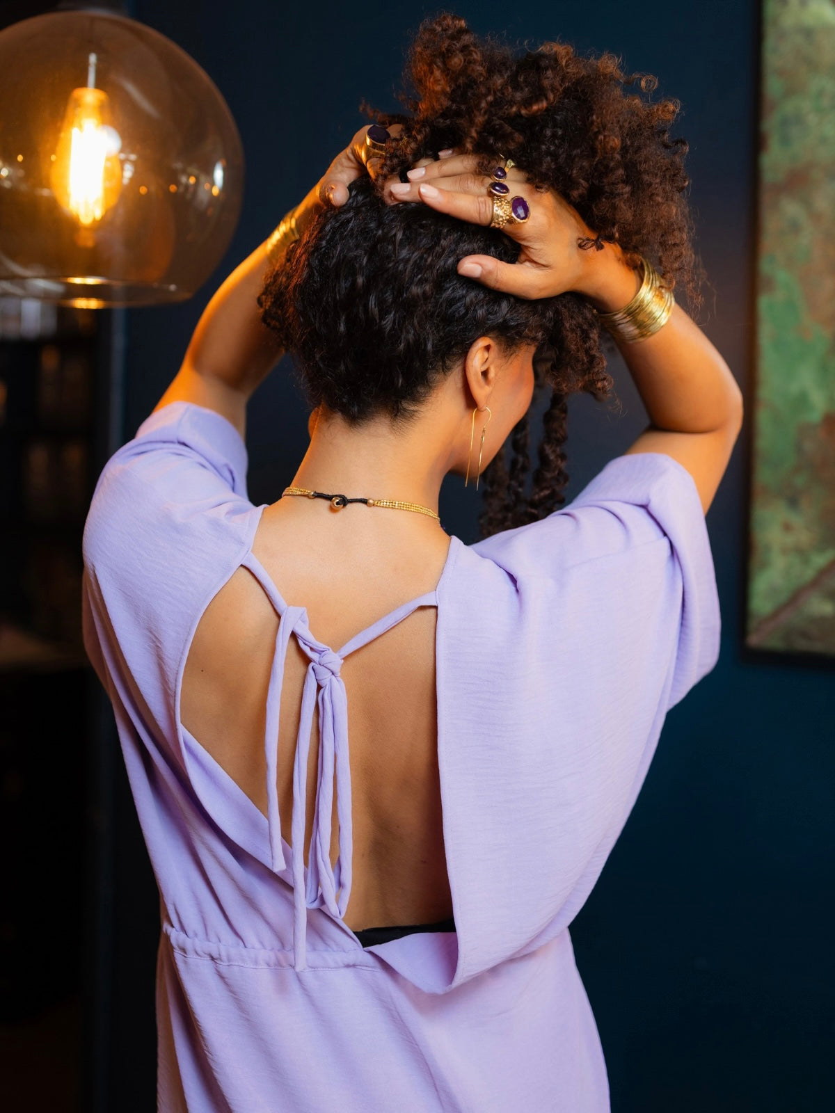 A woman with curly dark hair seen from behind, wearing a lavender-purple short-sleeve top with an open back and tie detail. She is standing indoors against a dark blue wall, with a glowing glass pendant light visible on the left.