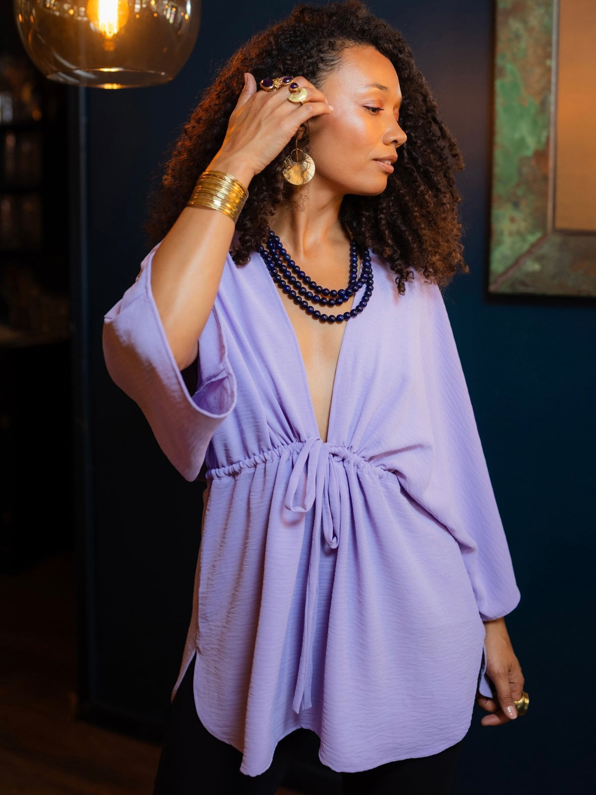 A woman wearing a lavender purple wrap-style tunic top with three-quarter sleeves and a drawstring waist, styled with layered dark beaded necklaces and gold jewelry in an indoor setting.