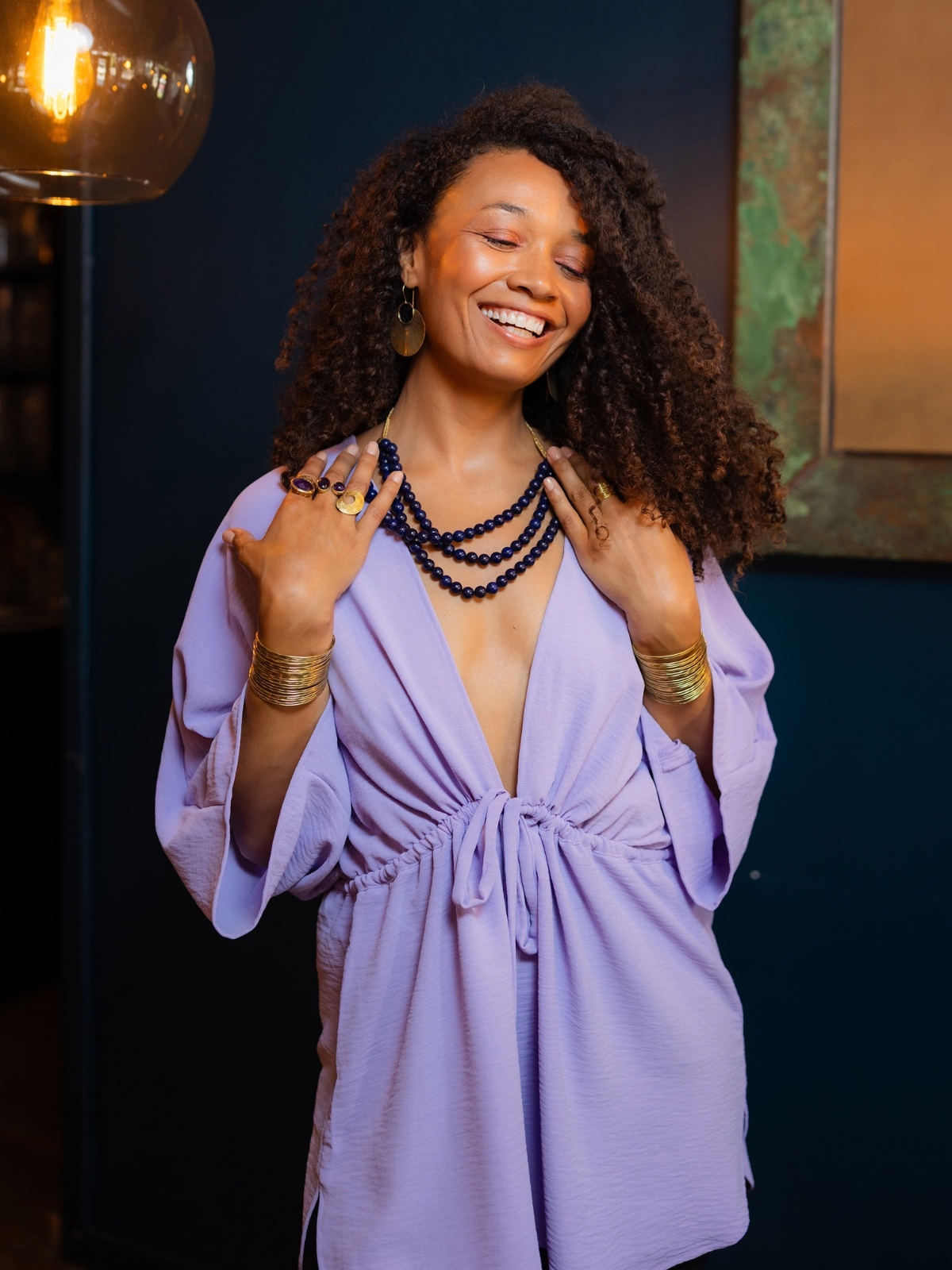 A woman with curly hair wearing a lavender purple wrap dress, layered black beaded necklaces, and gold bracelets, posing in a dimly lit indoor setting with a warm hanging light bulb visible in the background.