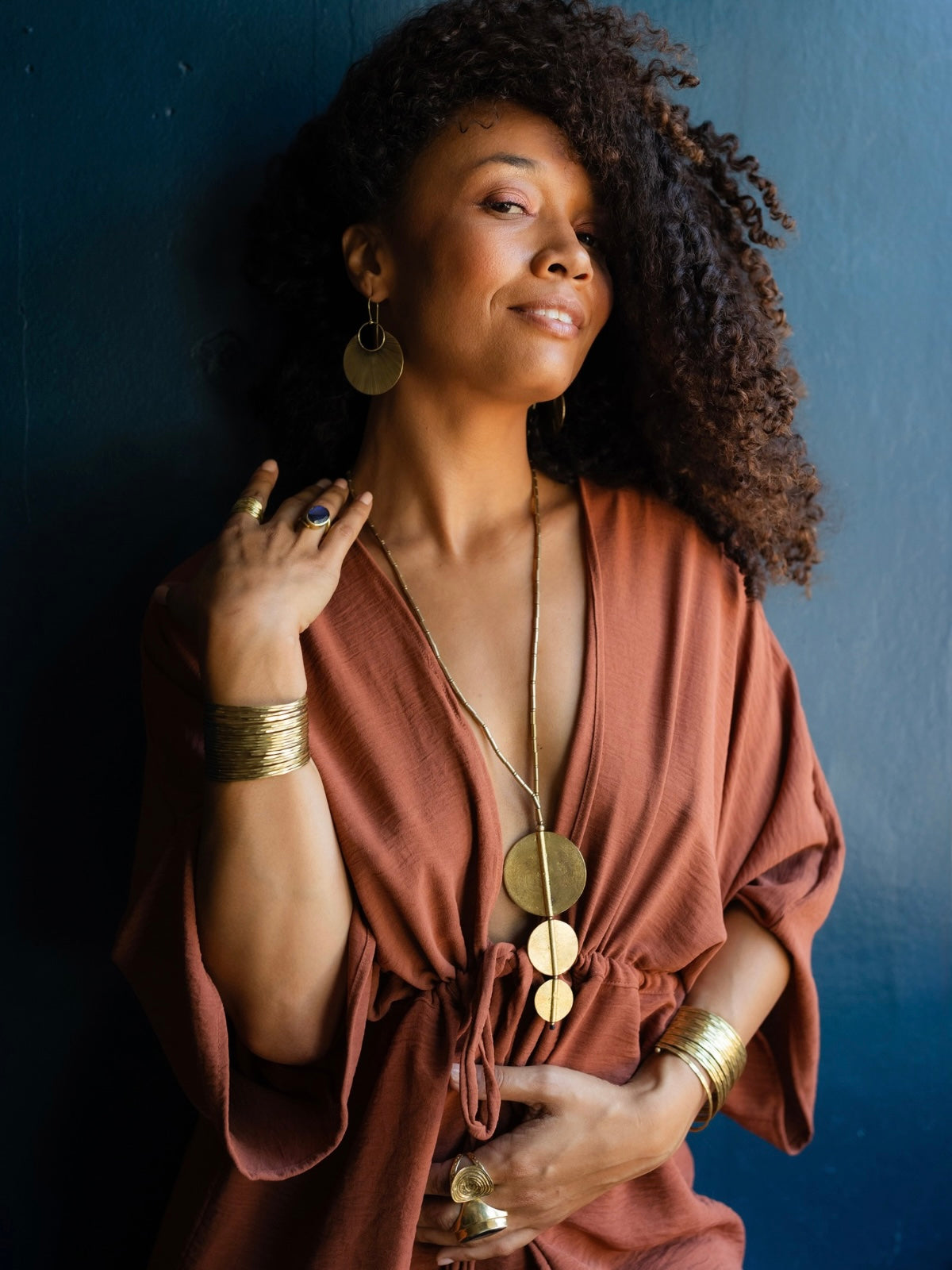 A woman with curly dark hair wearing a rust-brown draped top and gold jewelry, posing against a dark blue wall.