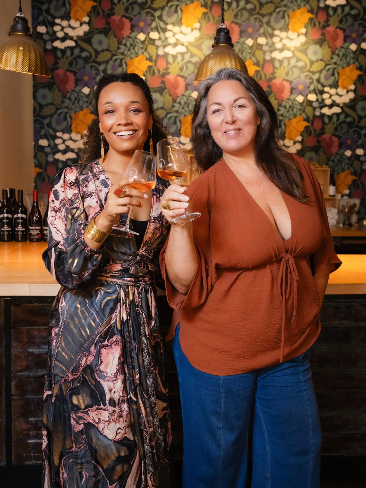 Two women posing together in a warmly lit bar or restaurant setting, each holding a wine glass. The woman on the left wears a long-sleeve floral print dress, and the woman on the right wears a rust-brown V-neck top and blue jeans.
