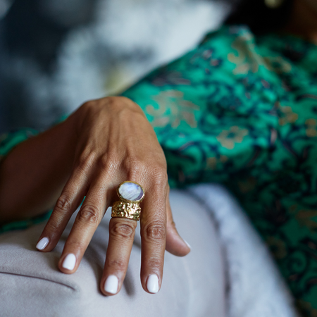 Hand wearing a gold ring with a large moonstone, against a blurred background.