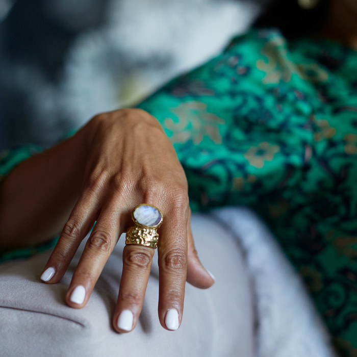 Hand wearing a gold ring with a large moonstone, against a blurred background.