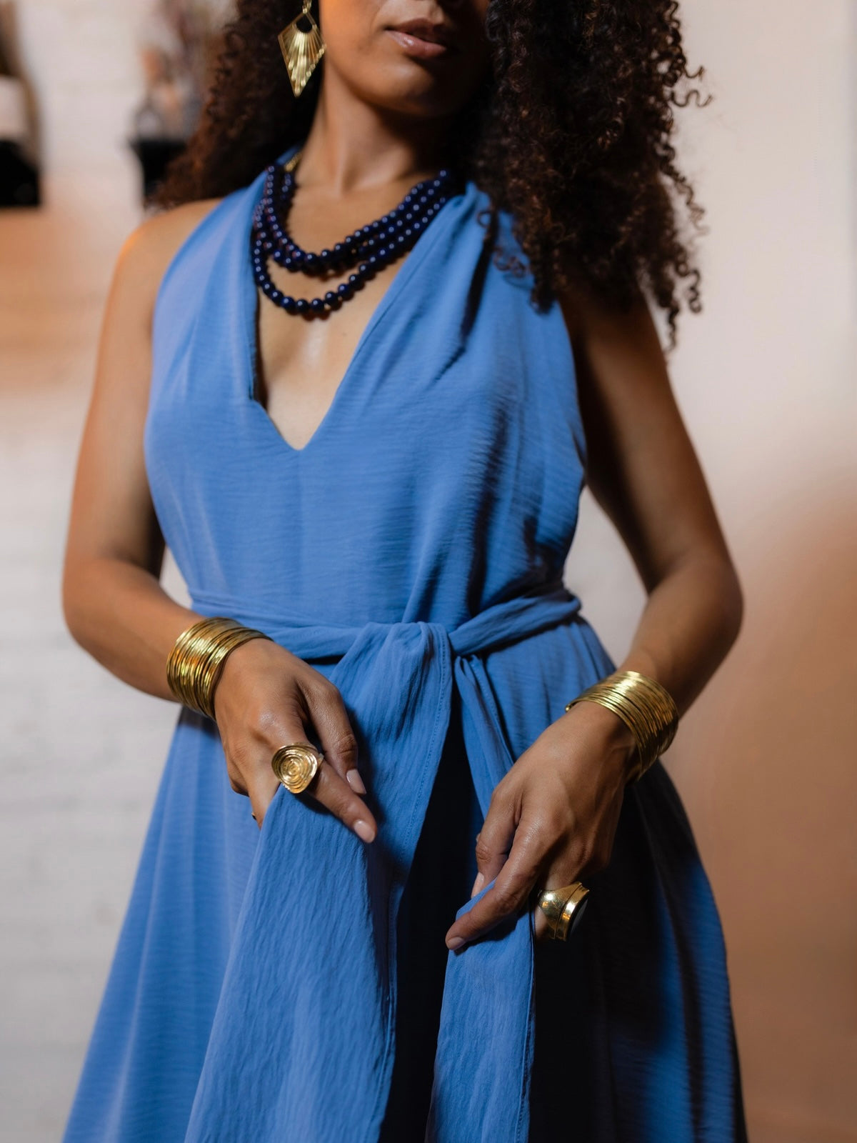 A woman wearing a sleeveless blue wrap-style dress with a deep V-neck, styled with layered dark blue beaded necklaces, gold bangles, and rings.