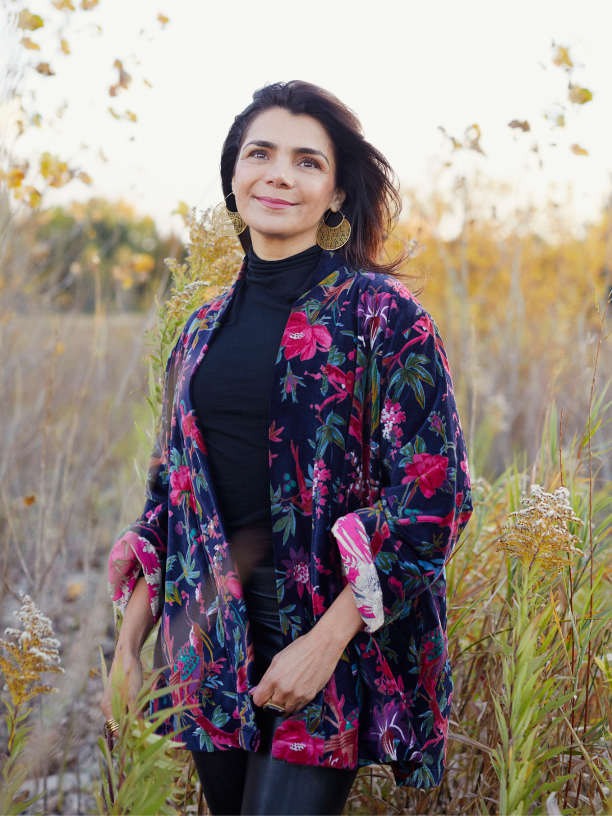 A woman standing outdoors in a field of dry grass, wearing a dark navy floral kimono jacket with pink and red flowers over a black turtleneck top.