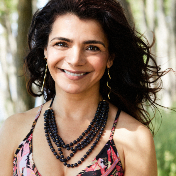 A smiling woman with dark hair wearing a floral halter top, layered black beaded necklaces, and gold swirl earrings, photographed outdoors against a soft blurred background of light-coloured trees.