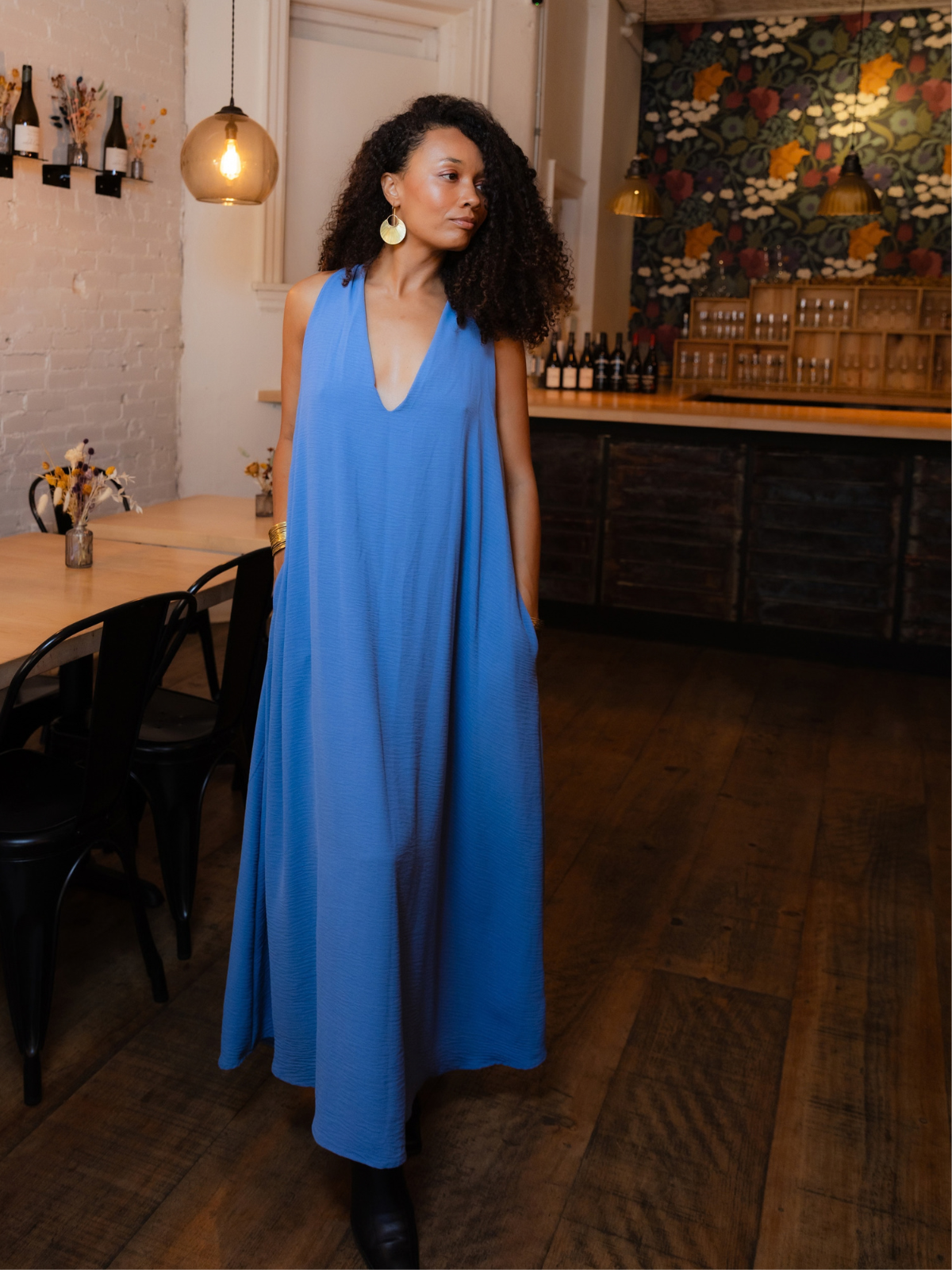 A woman with curly dark hair wearing a sleeveless, deep V-neck blue maxi dress with pockets, standing in a rustic restaurant or bar with wooden floors, a dining table, and a patterned accent wall.