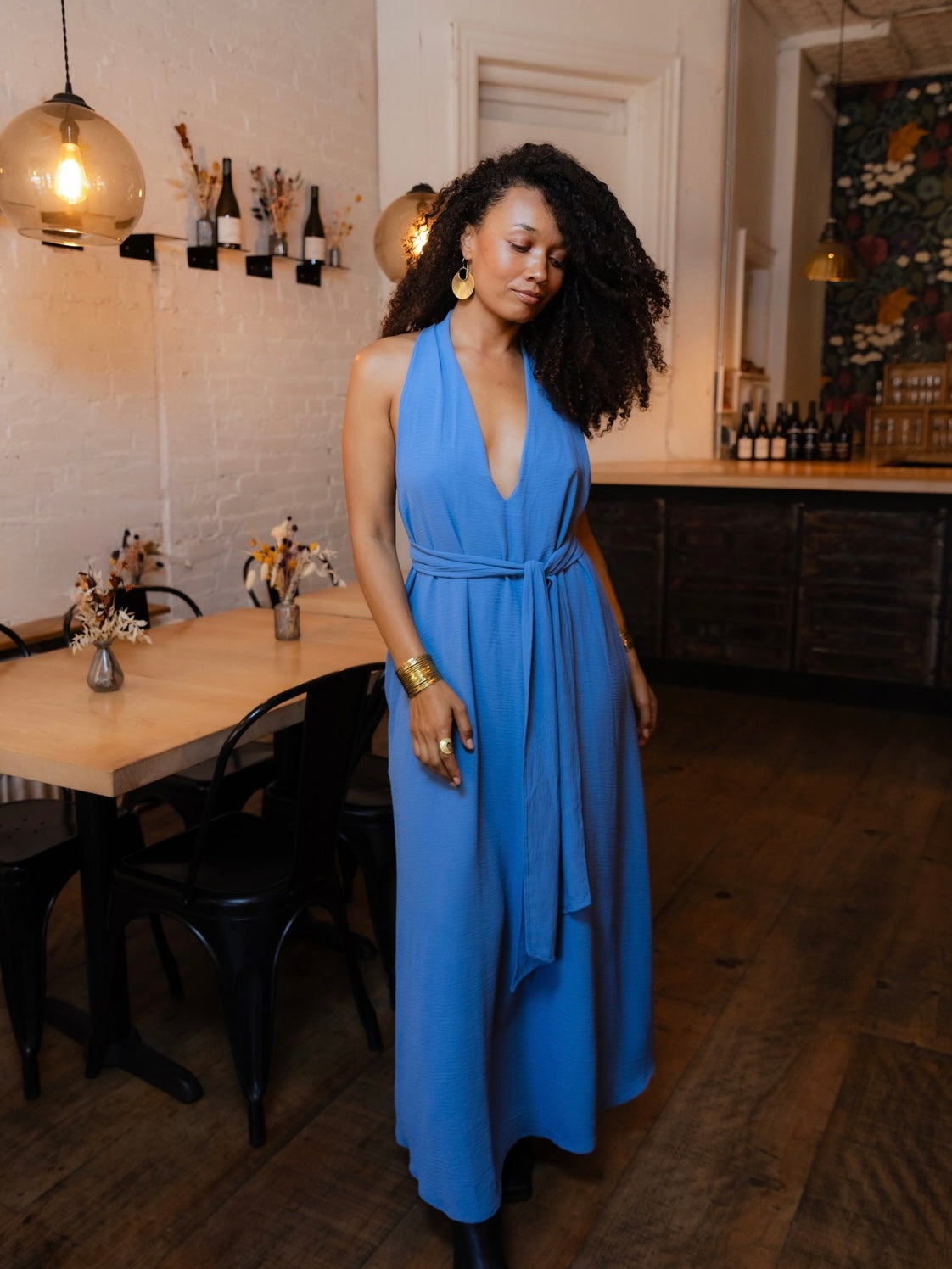 A woman with curly black hair wearing a bright blue halter-neck maxi dress with a tie waist, standing in a rustic restaurant or cafe with wooden floors, a table, and pendant lights.