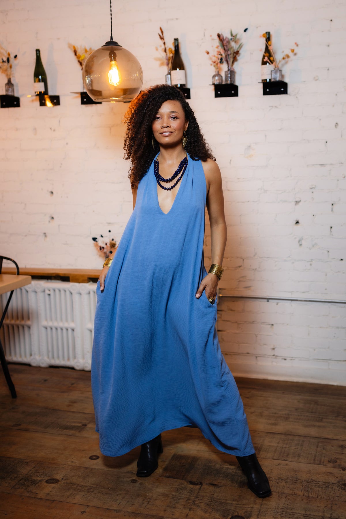 A woman with curly hair wearing a sleeveless, V-neck blue maxi dress with pockets, standing in a room with a white brick wall and wooden floor.