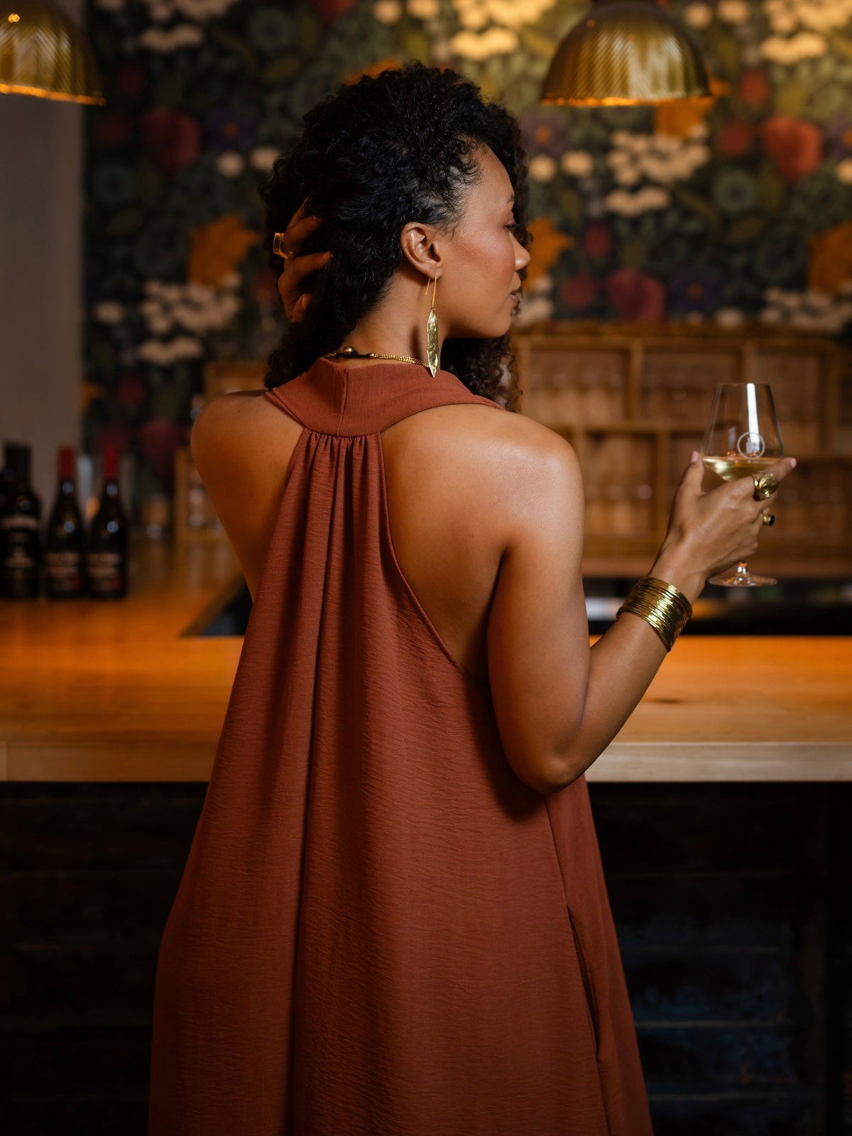 A woman stands at a bar, shown from the back and side, wearing a rust-brown sleeveless draped dress and holding a glass of white wine. The background features a dark floral wallpaper and warm ambient lighting.