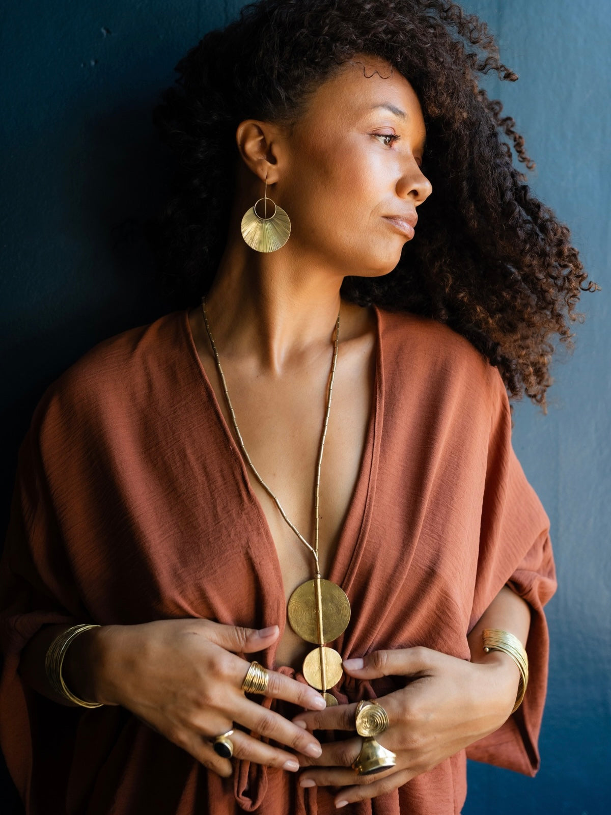 A woman with curly dark hair wearing a rust-colored top and multiple gold-toned jewelry pieces, including a long disc pendant necklace, hoop earrings, and rings, posing against a dark blue wall.