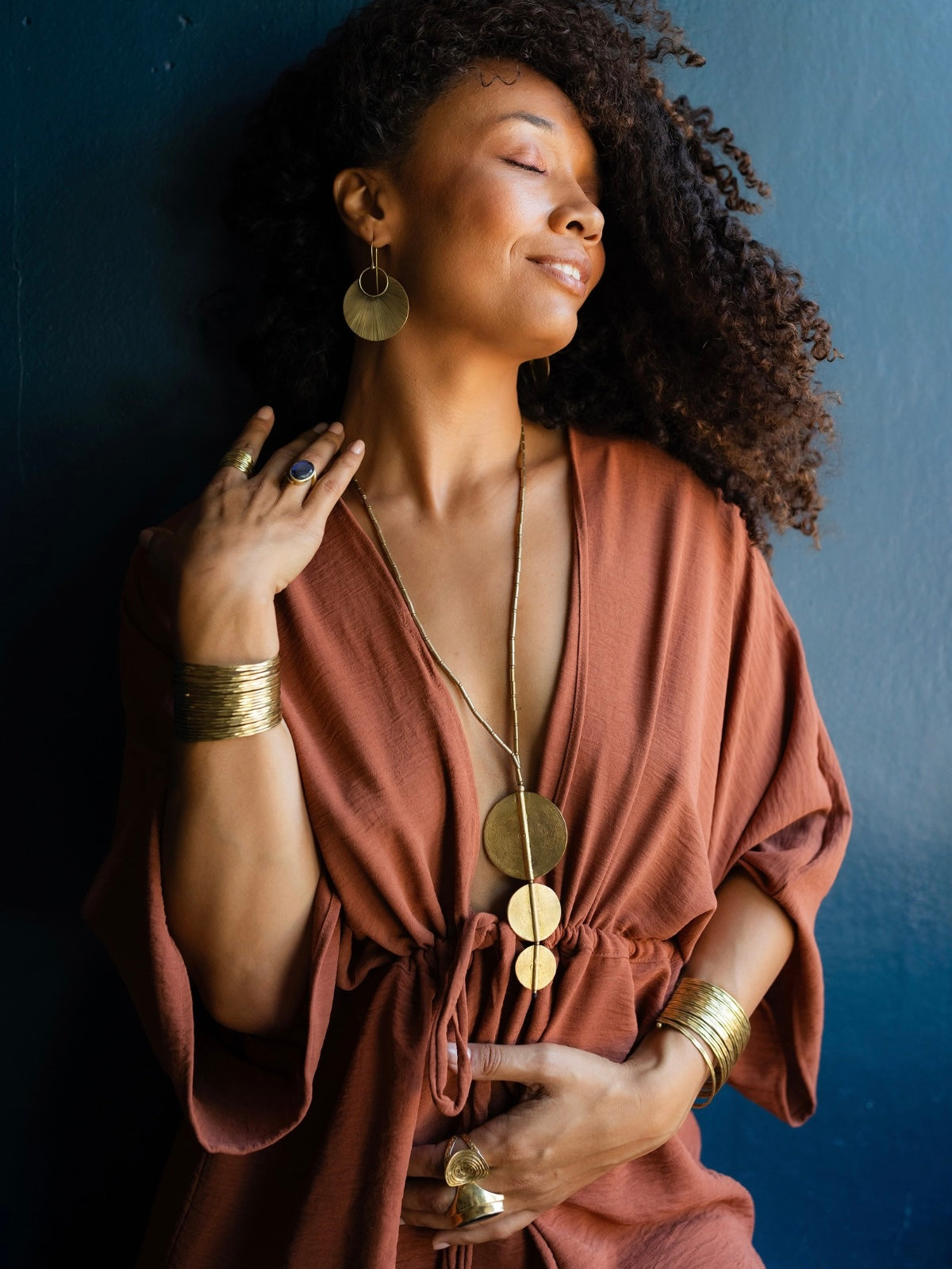 A woman with curly dark hair wearing a rust-brown wrap dress and gold jewelry, posing against a dark blue wall.