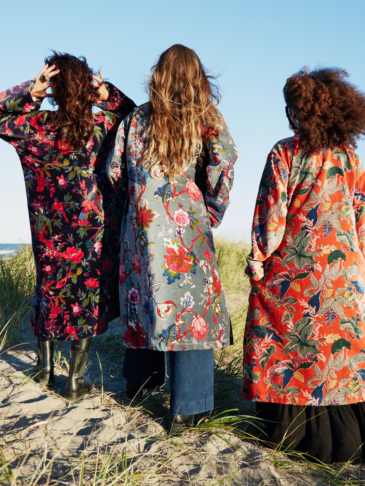 Three women standing on a sandy beach dune, viewed from behind, wearing long, loose floral-patterned coats in different colors and prints against a blue sky.