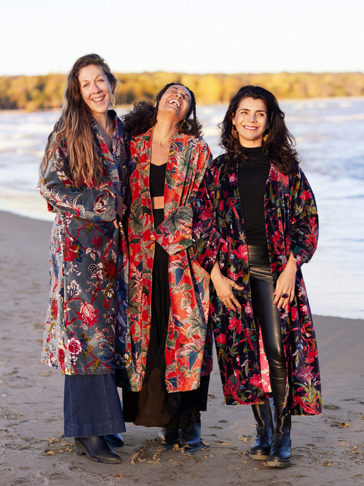 Three women standing together on a beach wearing long, open-front floral robes in different colorways. The left robe is blue-gray with red and pink floral patterns, the center robe is red with orange and green tropical floral patterns, and the right robe is black with red and pink floral patterns.